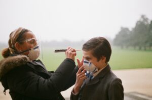 A makeup artist wearing a COVID mask and shield applies makeup to a child actor during a film shoot.